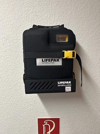 AED in schwarzer Tragetasche hinter der zweiten Tür links an der Wand über dem Feuerlöscher AED in schwarzer Tragetasche hinter der zweiten Tür links an der Wand über dem Feuerlöscher