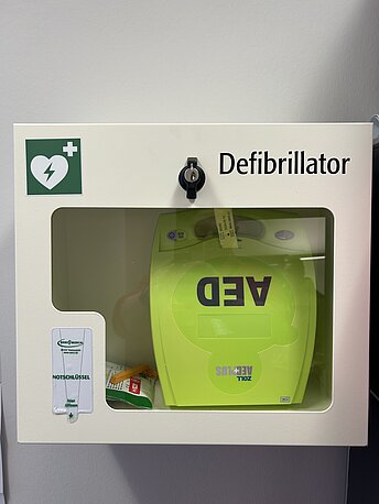 AED im weißen Wandkasten im Kopierraum hinter dem Empfang. Bitte am Empfang fragen!