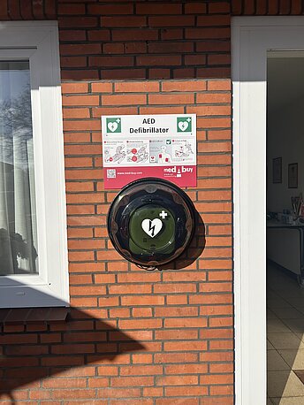 Öffentlicher AED im runden Wandkasten neben dem Eingang zum Dorfgemeinschaftshaus Öffentlicher AED im runden Wandkasten neben dem Eingang zum Dorfgemeinschaftshaus
