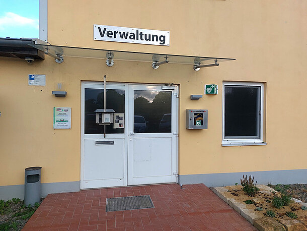 Öffentlicher AED im eckigen, silbernen Wandkasten rechts neben dem Haupteingang (Verwaltung) der Firma Happy Food