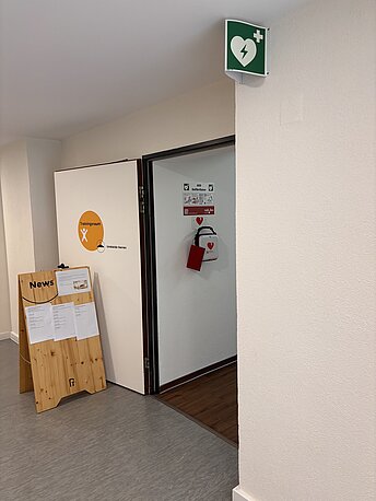 AED in weißer Transportasche links an der Wand direkt hinter der Tür.