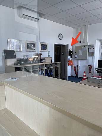 AED im Raum hinter dem Empfangstresen, hinter der Tür zum Nebenraum in schwarzer Tasche an der Wand.