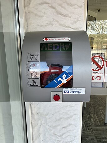 Öffentlicher AED im silbernen Wandkasten neben dem Haupteingang zur Volksbank Asendorf
