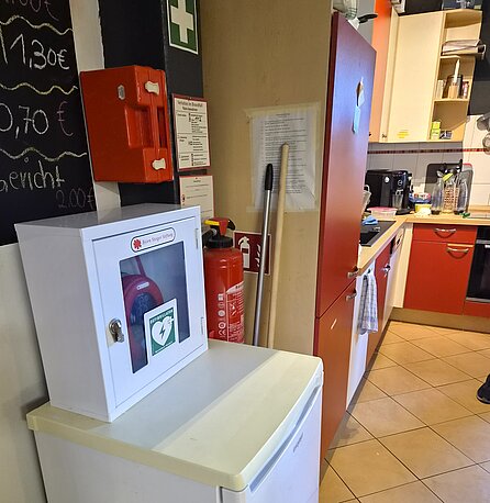AED im weißen Kasten auf dem Kühlschrank bei der Theke/Küche (Offenes Jugendcafé)
