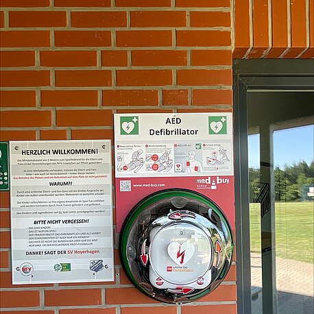 Öffentlicher AED am Funktionsgebäude auf der Sportplatzseite gegenüber dem Klettergerüst in der Gebäudenische (Eingang zu den Umkleiden) im runden Wandkasten.