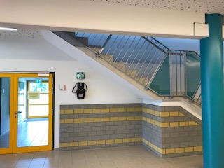 AED wandmontiert unter der Treppe, rechts von der Glastür zum Sportplatz AED wandmontiert unter der Treppe, rechts von der Glastür zum Sportplatz